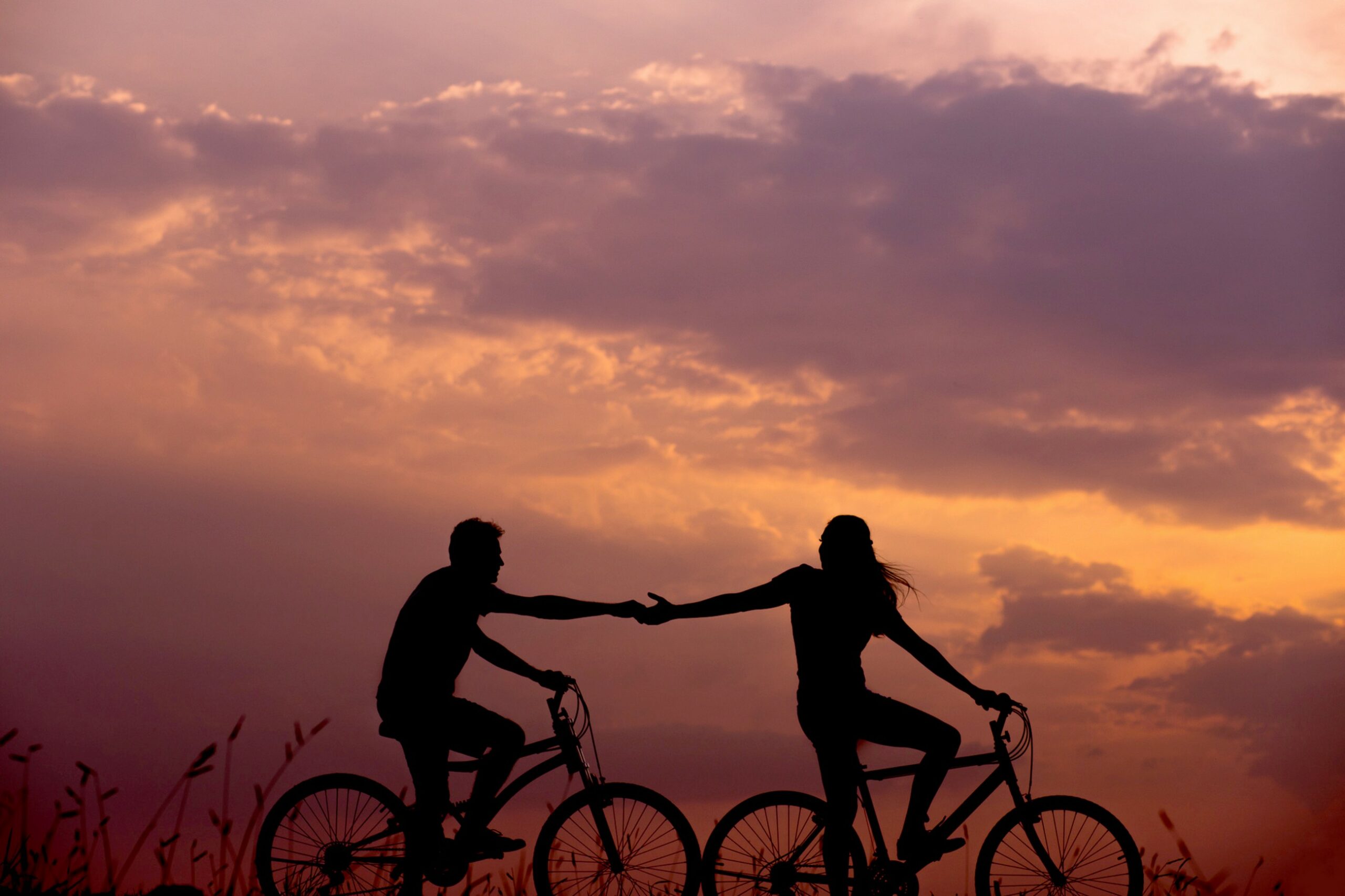 Sunset, two bike riders silhouetted in the foreground. The lead lady is reching back and grasping hands with the male. Romantic.